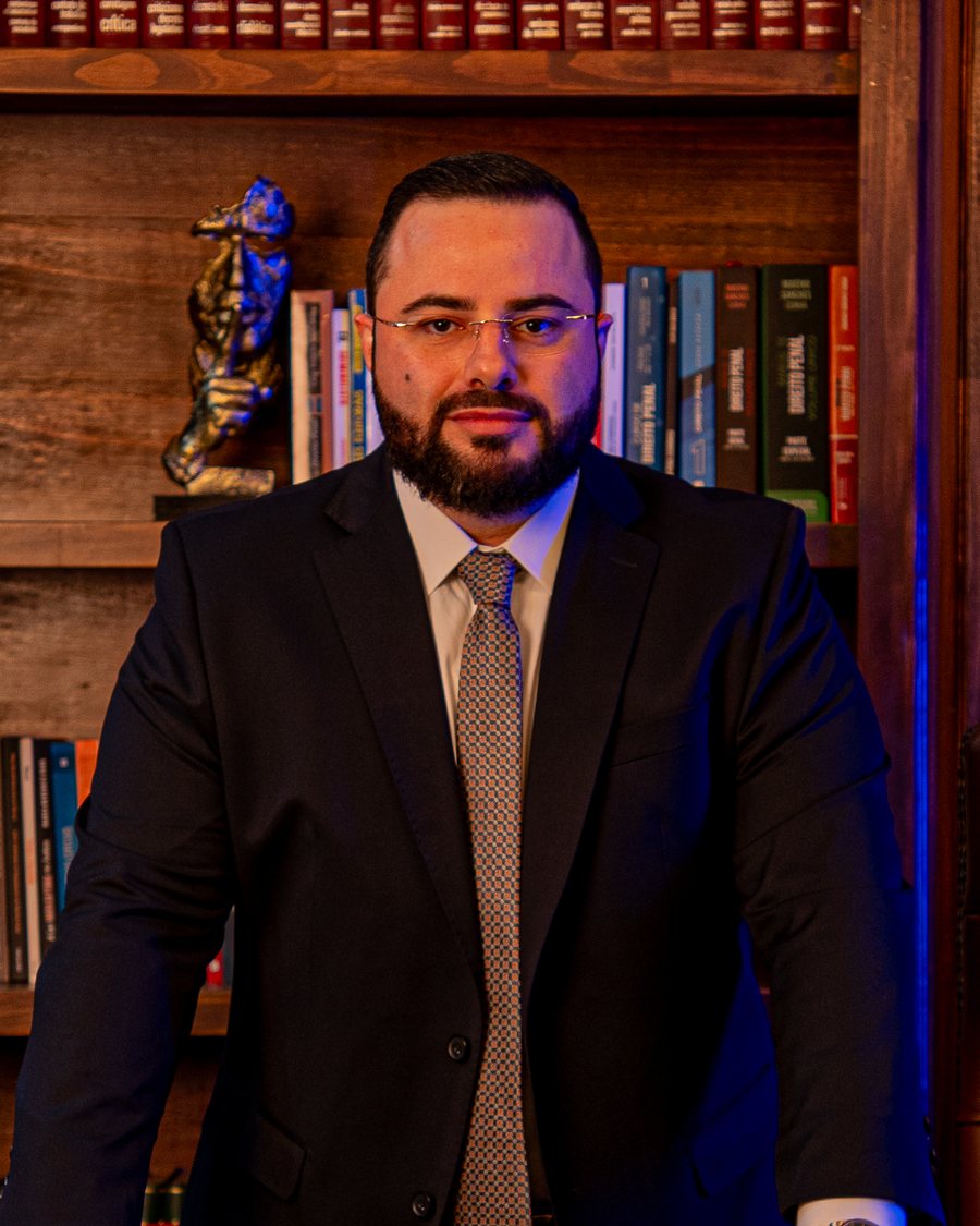 Retrato do advogado criminalista Daniel Broeto Maia, de terno escuro, apoiado em mesa diante de estante de livros jurídicos